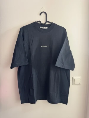 Svart t-shirt Acne Studios - Svart t-shirt från Acne Studios med vit logotyp på bröstet. Modellen är loose fit med korta ärmar och rund hals. Materialet är mjuk bomull som känns skönt mot huden. Perfekt för dig som gillar minimalistisk och stilren design.