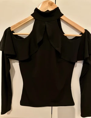 Otroligt vacker topp med volang och cut-out från Nelly - Säljer en snygg svart topp från NLY med lång ärm och snygga volangdetaljer över axlarna. Haft på mig bara 1 gång. Toppen har en cool cut-out design vid axlarna och hög krage med knappar i nacken. Materialet är mjukt och stretchigt, perfekt för dig som gillar statement-plagg.