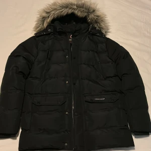 Svart Canada Goose Jacka - Säljer en svart dunjacka från Canada Goose i storlek L. Jackan har huva, flera fickor med lock och tryckknappar samt klassisk Canada Goose-logga på ärmen. Perfekt för kalla vinterdagar och riktigt snygg streetstil.