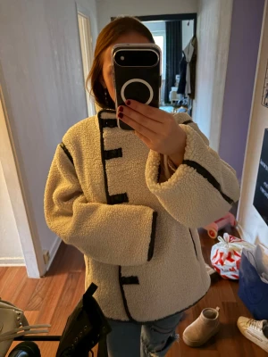 Beige teddyjacka med svarta detaljer - Säljer en beige teddyjacka med svarta kantband och dekorativa knappar framtill. Jackan har en boxig, oversized passform och är gjord i mjukt, fluffigt material som håller dig varm. Perfekt för dig som vill ha en trendig och mysig look.