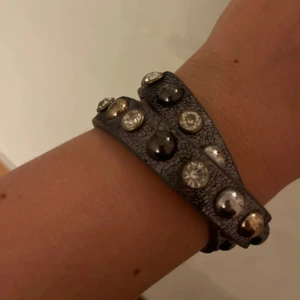 Svart läderarmband med nitar och stenar - Coolt svart läderarmband som snurras två varv runt handleden. Prytt med blanka nitar i silver och guld samt glittrande stenar. Perfekt accessoar för att ge outfiten lite extra edge och stil.