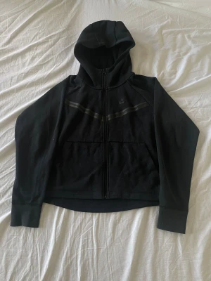 Nike Tech Fleece  - Svart hoodie från Nike med hel dragkedja och huva. Tröjan har två fickor framtill och ett diskret Nike-logga på bröstet. Snygga svarta ränder går över bröstet. Tillverkad i mjuk bomullsmix, perfekt för chill eller träning.