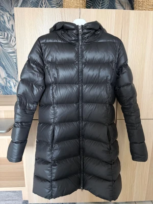 Svart lång dunjacka från Colmar - Snygg svart lång dunjacka från Colmar med huva och quiltad design. Jackan har dragkedja framtill och logga på ärmen. Perfekt för kalla dagar och ger en clean, stilren look. Materialet är glansigt och jackan är lätt vadderad för extra värme.