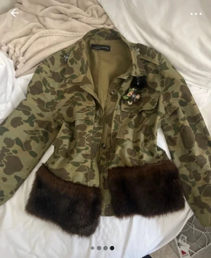 Militärmönstrad jacka med pälsdetalj Zara - Unik camouflagejacka från Zara med bruna pälsdetaljer längst ner och broderade blommor på bröstet. Jackan har knappar framtill, två stora bröstfickor och coola detaljer som ger en edgy vibe. Perfekt för dig som vill sticka ut med din stil.