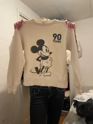Beige Disney Mickey Mouse tröja - Beige sweatshirt från Stradivarius x Disney med stor Mickey Mouse-tryck och texten '90 Years' framtill. Långärmad modell med ribbade muddar och rund hals. Mjuk insida och tryckt text längs ena ärmen. Perfekt för dig som älskar Disney och vill ha en chill look.