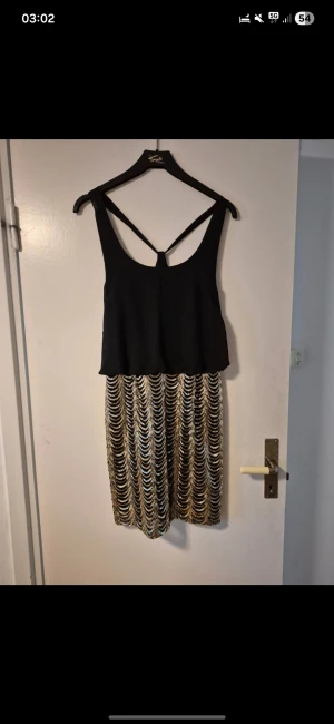 Svart och guld paljettklänning - Supersnygg kort klänning med svart överdel och glittrig kjol i guld paljetter. Kjolen har ett vågigt mönster och överdelen har breda axelband med snygg ryggdetalj. Perfekt för dig som vill sticka ut med en festlig look.