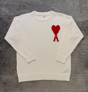 Vit stickad tröja från Ami Paris - Vit stickad tröja från Ami Paris med röd hjärt- och A-logga på bröstet. Rund halsringning och ribbade muddar vid ärmslut och nederkant. Tröjan har en clean och stilren look som passar perfekt till jeans eller chinos. Materialet är mjukt och skönt.
