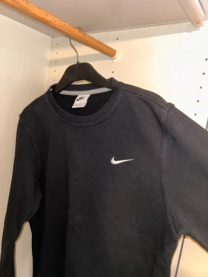 Svart sweatshirt från Nike - Svart sweatshirt från Nike med klassisk rund hals och broderad vit Nike-logga på bröstet. Tröjan har lång ärm och är gjord i mjuk bomullsmix som är skön mot huden. Perfekt för chill dagar eller när du vill ha en enkel och stilren look.
