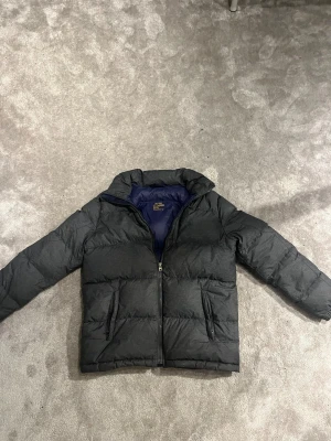 Mörkgrå pufferjacka Polo Ralph Lauren  - Snygg mörkgrå pufferjacka från Polo Ralph Lauren i storlek XS (XL barn) . Jackan har en quiltad design, dragkedja framtill och två fickor med zip. Insidan är marinblå och jackan har broderad logga på bröstet. Perfekt för kalla dagar och riktigt chill stil. Luva saknas tyvärr men fortfarande tvär snygg på! Vid funderingar hör av er! 