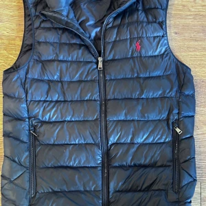 Ralph Lauren väst - Snygg marinblå dunväst från Polo Ralph Lauren i storlek XS men skulle säga att den sitter som en S. Västen har quiltad design, dragkedja framtill och två fickor med dragkedja. Klassisk röd logga broderad på bröstet och hög krage. Västen har ett litet hål på ryggen(se bild 4) men inget som syns vid användnig.