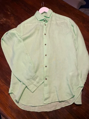 Grön linneskjorta från Camicissima - Fräsch grön skjorta från Camicissima i linnematerial, perfekt för varma dagar. Skjortan har klassisk krage, bruna knappar och lång ärm. Comfort fit och snygg enkel design som ger en chill vibe. Skjortan är bara använd 1-2 gånger 