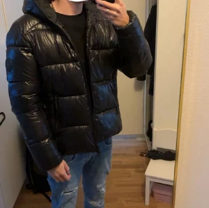 Svart pufferjacka från Save The Duck - Säljer en svart pufferjacka från Save The Duck med glansig finish och huva. Jackan är vadderad med syntetisk Plumtech-fyllning och har yttertyg i nylon. Utrustad med dragkedja framtill och två sidofickor. Perfekt för kalla dagar och riktigt snygg streetstil. Nypris runt 3000kr. Strlk S. 