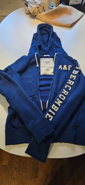 Blå hoodie Abercrombie & Fitch L - Snygg marinblå hoodie från Abercrombie & Fitch i storlek Large. Tröjan har huva med snörning, dragkedja och ribbade muddar. Stor vit logotyp på ärmen och A&F på bröstet. Tillverkad i mjuk bomull och polyester för skön känsla.