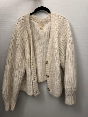 Beige stickad kofta från H&M alpacka blend - Mysig beige stickad kofta från H&M med grov ribbstickning och stora knappar framtill. Koftan har långärmade ärmar och en avslappnad passform som ger en chill vibe. Perfekt att slänga på sig när det är lite kyligt ute.