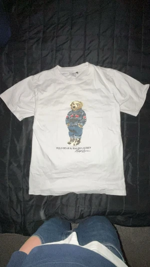 Vit Polo Bear t-shirt från Ralph Lauren - Vit t-shirt från Ralph Lauren med ikoniskt Polo Bear-tryck på bröstet. Björnen har en stickad tröja med rött och blått mönster, jeans och boots. T-shirten har rund hals och korta ärmar, tillverkad i mjuk bomull. Perfekt för dig som gillar klassisk streetstyle.
