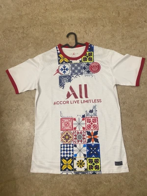 PSG x Jordan vit fotbollströja M - Unik PSG x Jordan fotbollströja i vitt med röda detaljer på krage och ärmslut. Framsidan har färgglada grafiska mönster i blått, gult och rött, samt klubbmärke och Jordan-logga. Tillverkad i lätt polyester som andas. Perfekt för dig som vill sticka ut på planen.