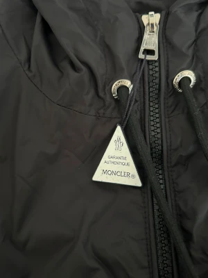 Svart vindjacka från Moncler med huva - Svart vindjacka från Moncler med huva och coola detaljer som snörning och dragkedja framtill. Jackan har Moncler-logga på ärmen och snygga ränder i rött, vitt och blått på huvan. Perfekt för dig som gillar stilrena och sportiga plagg.