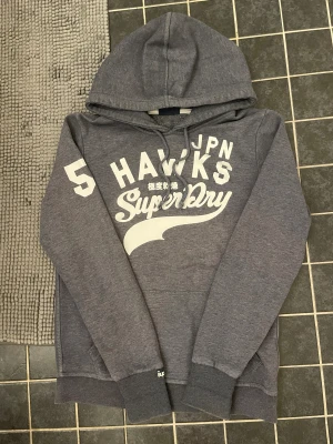 Grå hoodie Superdry med tryck - Snygg grå hoodie från Superdry i storlek Large med stort vitt tryck på bröstet och ärmen. Tröjan har huva med snörning och ribbade muddar vid ärmslut och nederkant. Perfekt för chill dagar och streetstyle. Det står storlek L men passar nån runt 165-175.