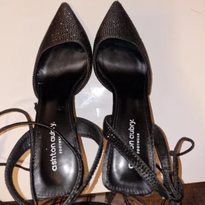 Svarta glittriga pumps med snörning - Svarta pumps från Ashton Aubry med spetsig tå och hög klack. Skorna har glittriga detaljer över hela ovandelen och tunna snören som knyts runt ankeln. Snygg och elegant design med platt sula framtill och stilren finish. Perfekt för dig som gillar att sticka ut.