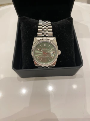 Seiko Datejust med grön urtavla - Säljer en stilren Seiko Oyster Perpetual Datejust med grön urtavla och silverfärgat armband i metall. Klockan har klassisk rund form, räfflad bezel och datumvisning med förstoring. Länken är polerad och har en snygg låsning. Automatisk rörelse syns genom bakboetten.