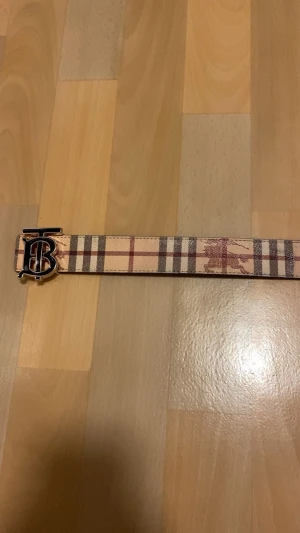 Burberry bälte med rutigt mönster - Säljer ett snyggt Burberry bälte med klassiskt rutigt mönster i beige, brun och röd. Bältet har en metallspänne med TB-logga och är tillverkat i skinn. Perfekt accessoar för att lyfta din outfit med en lyxig touch.