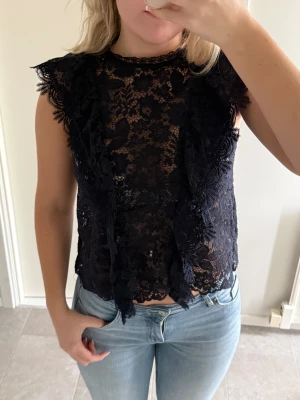 Svart spetsblus med volangdetaljer - Säljer en mörkblå blus i genombruten spets med blommigt mönster. Blusen har volangdetaljer och man knäpper i nacken