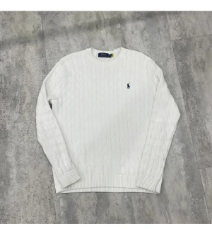 Vit stickad tröja från Polo Ralph Lauren - Vit kabelstickad tröja från Polo Ralph Lauren med klassisk rund hals och blå broderad logga på bröstet. Tröjan har ribbade muddar vid ärmslut och nederkant, och är långärmad. Perfekt för dig som gillar stilrena och tidlösa plagg.