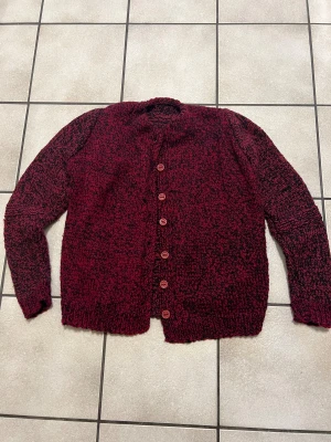 Mörkröd stickad ull kofta med knappar storlek S - Säljer en chunky stickad kofta i mörkröd och svart melerad färg. Koftan har långa ärmar, rund hals och sex röda knappar framtill. Grovstickad look som ger en cool och avslappnad vibe, perfekt för kyliga dagar.