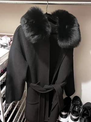 Svart kappa med pälsdetalj - Snygg svart kappa med bred krage i fluffig pälsimitation och knytbälte i midjan. Jackan har en stilren och rak passform, perfekt för dig som vill ha en elegant look med extra attityd. Pälsdetaljen ger en lyxig vibe och jackan är helsvart.