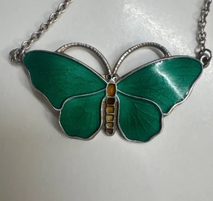 Grönt fjärilshalsband från Opro - Unikt halsband i form av en fjäril från Opro, tillverkat i 925 sterling silver. Fjärilen har vingar i en snygg grön färg med detaljer i gult och brunt på kroppen. Kedjan är tunn och silverfärgad, perfekt för dig som gillar naturinspirerade smycken.