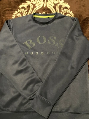 Svart sweatshirt från Hugo Boss - Svart långärmad sweatshirt från Hugo Boss med stor BOSS-logga framtill. Tröjan har rund hals och är tillverkad i mjuk polyester som ger en clean och modern look. Perfekt för dig som gillar stilrena och klassiska märkesplagg.