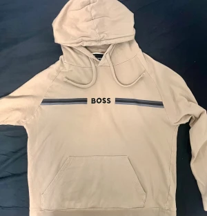 Boss hoodie/tröja - Snygg beige hoodie från BOSS med svart tryck och två mörkgrå ränder över bröstet. Tröjan har huva med snörning och en stor magficka framtill. Perfekt för dig som gillar stilrena och enkla plagg med en touch av street. Köpt för 1200kr från Boozt. Den e helt fin och har inga problem.