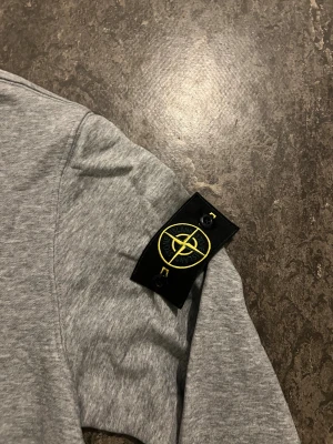 Grå sweatshirt från Stone Island - Snygg grå sweatshirt från Stone Island med klassisk rund hals och ribbade muddar. Tröjan har den ikoniska Stone Island-patchen på vänster ärm och är tillverkad i mjuk bomull. Perfekt för dig som gillar stilrena och sportiga plagg.