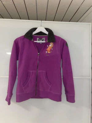 Söt fleecejacka CRW med hästtryck - Lila fleecejacka från CRW med mysig mörkgrå krage och supergulligt tryck av en tecknad häst på bröstet. Jackan är i barnstorlek 146/152 från Hööks✨  Eftersom jag tyckte att den var för fin för att ha i skolan, användes den ca 2/3 ggr.   I övrigt är det inga tecken på användning.