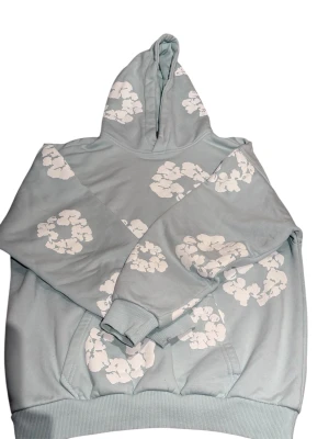 DENIM TEARS HOODIE LJUSBLÅ - Snygg ljusblå hoodie med huva och vita blommönster över hela tröjan. Mjuk bomullskänsla, ribbade muddar och praktisk ficka framtill. Perfekt för dig som gillar chill och stilren design med en twist.