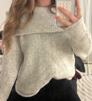 Ljusgrå stickad tröja med bred krage - Mysig ljusgrå stickad tröja med bred, ribbad krage som går över axlarna. Tröjan har långärmade ärmar och en loose passform, perfekt för att hålla sig varm under kyliga dagar. Enkel och stilren design som är lätt att matcha med olika outfits.