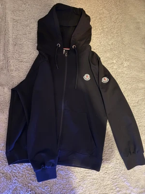 Svart Moncler hoodie med dragkedja - Svart hoodie från Moncler med klassiska logopatchar på bröstet och ärmen. Tröjan har huva med snörning, hel dragkedja och rymliga fickor framtill. Tillverkad i mjuk bomull, perfekt för chill dagar. Snygg och enkel design som funkar till allt.