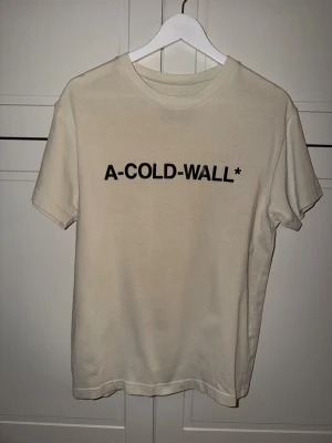 Beige t-shirt från A-COLD-WALL* - Snygg beige t-shirt från A-COLD-WALL* med svart logotyptryck över bröstet. Klassisk rund halsringning och korta ärmar. Tillverkad i mjuk bomull för en skön känsla och clean look.