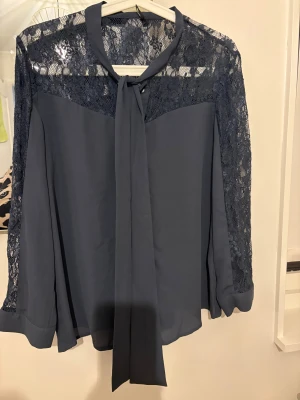 Mörkblå blus med spets från ONLY - Mörkblå blus från ONLY med långa ärmar, spetsdetaljer över axlar och ärmar samt knytband i halsen. Blusen har knappar framtill och är tillverkad i ett lätt, flowy material som ger en snygg siluett.