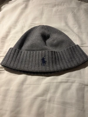 Grå merinoullmössa från Polo Ralph Lauren - Snygg grå mössa från Polo Ralph Lauren i mjuk och tvättbar merinoull. Klassisk ribbad kant och broderad marinblå logga framtill. Perfekt för dig som vill ha en stilren och varm mössa med premiumkänsla.