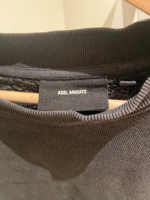Mörkgrå sweatshirt Axel Arigato - Säljer en mörkgrå sweatshirt från Axel Arigato med stora, broderade bokstäver på ryggen. Tröjan har en loose passform, rund hals och långa ärmar. Materialet är mjuk bomull och ger en chill vibe. Perfekt för dig som gillar streetwear och snygga detaljer.