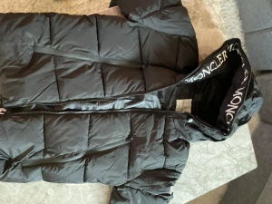 Svart Moncler Polyamide - Svart pufferjacka från Moncler med stor huva och vit logotyptext längs kanten. Jackan har dragkedja framtill, två sidofickor med dragkedja och quiltad design. Perfekt för kalla dagar och har en clean, modern look som sticker ut.
