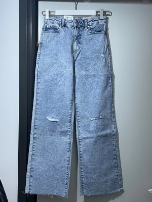 Ljusblå loose fit jeans från Mooi - Säljer ett par ljusblå jeans från Mooi med hög midja och loose fit. Byxorna har slitna detaljer framtill, raka ben och råa kanter vid bensluten. Klassiska femficksmodell med snygga bakfickor och vit patch med logga bak. Perfekt för en chill och trendig look.