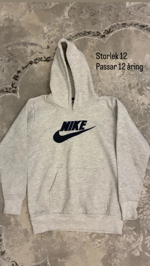 Grå hoodie från Nike med logga - Säljer en ljusgrå hoodie från Nike i storlek 12. Tröjan har en stor svart Nike-logga på bröstet, huva och en praktisk magficka. Perfekt för dig som gillar sportig och avslappnad stil.