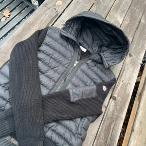 Byte - Byte Svart pufferjacka från Moncler med stickade ärmar och huva. Jackan har quiltad kropp, ribbade muddar och dragkedja framtill. Perfekt för kyliga dagar och har en snygg, sportig look med Moncler-logga på ärmen.