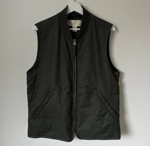 Mörkgrön nylonväst från ARKET, strl 48 - Mörkgrön stilren väst från ARKET i lätt nylonmaterial. Västen har två snedställda fickor med knappstängning och en kraftig dragkedja framtill. Ribbad krage ger en sportig touch. Perfekt att slänga över en hoodie eller t-shirt för extra lager. 5H6