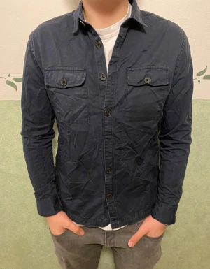 Mörkblå overshirt från River Island - Snygg mörkblå overshirt från River Island i storlek M. Skjortan har klassisk krage, två bröstfickor med knappstängning och knappar hela vägen framtill. Tillverkad i mjuk bomull som är skön mot huden. Perfekt att slänga på över en t-shirt för en chill look.