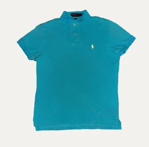 Ralph Lauren skjorta  - Turkos pikétröja från Polo Ralph Lauren med klassisk krage och två knappar framtill. Tröjan har det ikoniska broderade logot i vitt på bröstet och är i en slim fit-modell. Perfekt för dig som gillar stilren och sportig look. Pris kan diskuteras!