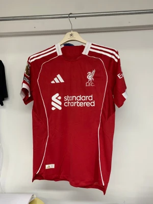 Liverpool FC röd matchtröja Isak #9 - Liverpool FCs officiella röd fotbollströja från Adidas med vita detaljer och tryck. Kortärmad, med Premier League-märke på ärmen och "No room for racism"-patch. Namn och nummer "ISAK 9" på ryggen. Tillverkad i ventilerande polyester.