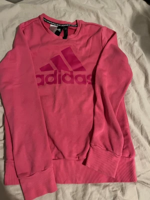 Rosa Adidas sweatshirt med logga - Säljer en snygg rosa sweatshirt från Adidas med stor logga framtill. Tröjan har rund hals, långa ärmar och breda muddar vid ärmslut och nederkant. Perfekt för dig som gillar sportig stil och vill sticka ut med färg.